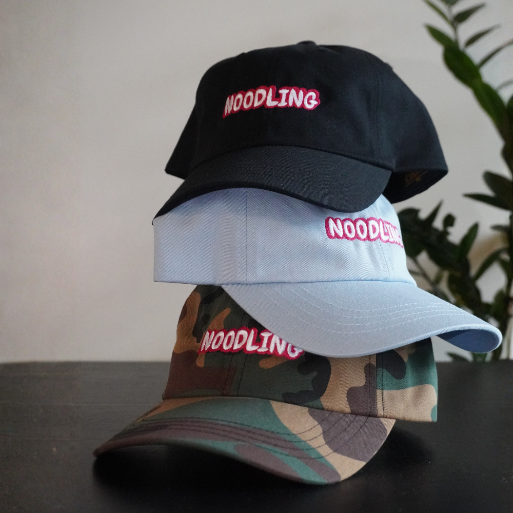 Three caps with 'NOODLING' logo stacked on a dark surface with a plant in the background.