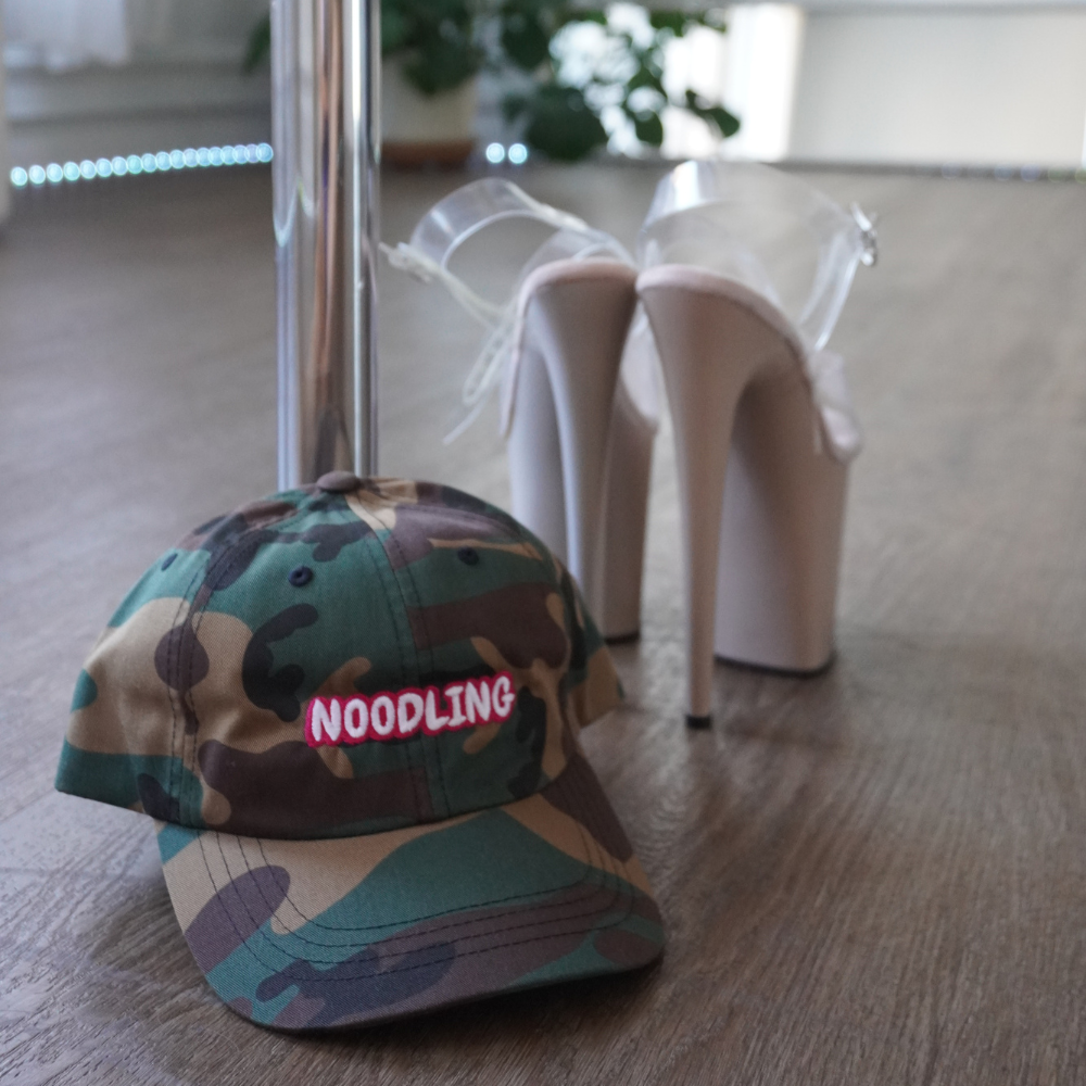 Camouflage cap with 'NOODLING' text and high-heeled shoes on a wooden floor.