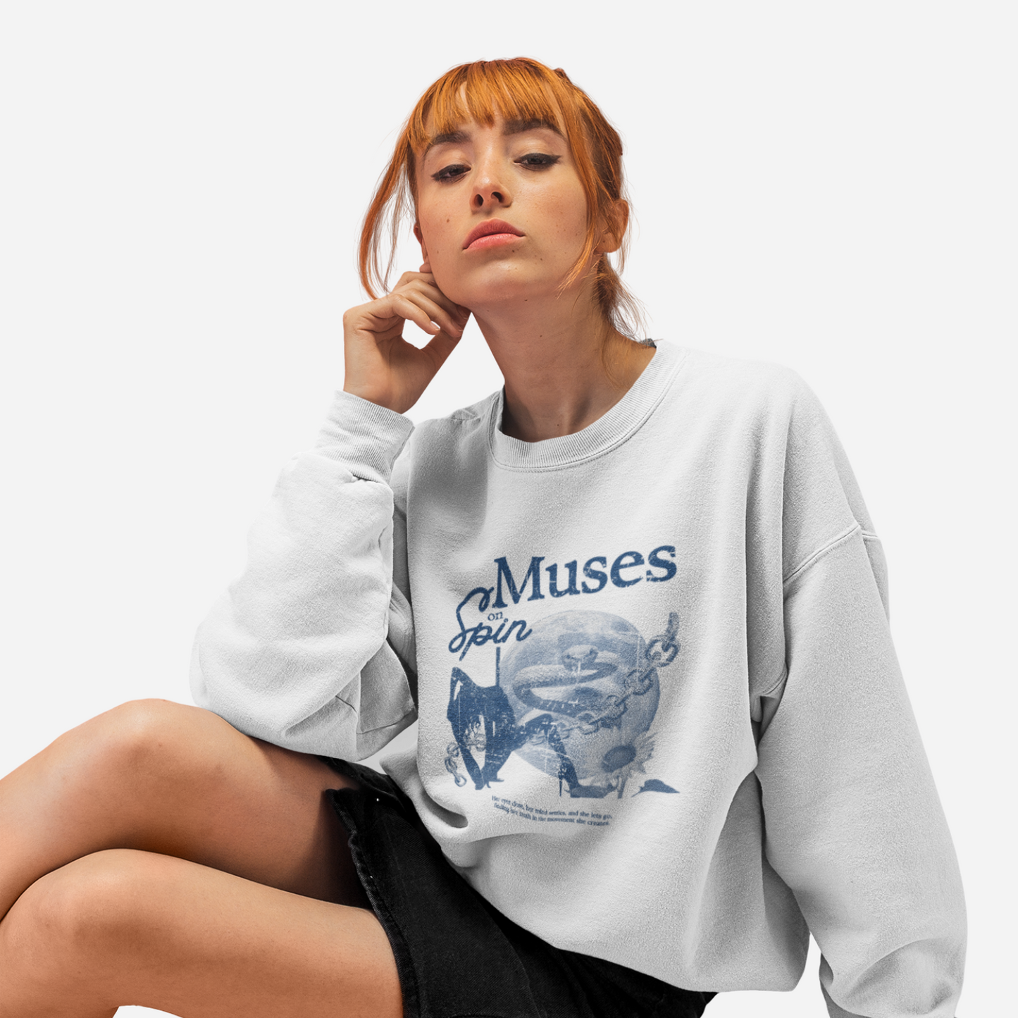 Woman sitting in front of a white background, wearing a white sweater with a blue graphic that includes a dancer, a moon, a snake, a chain, a flower and the text "Muses on Spin".