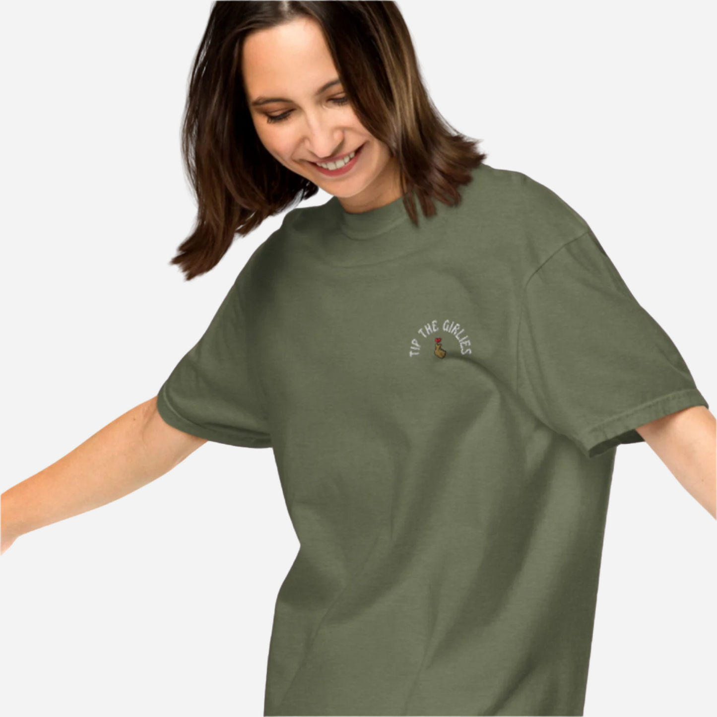 Woman in front of a white background wearing a loose green tshirt with the words "Tip the Girlies" embroidered on the left chest area.