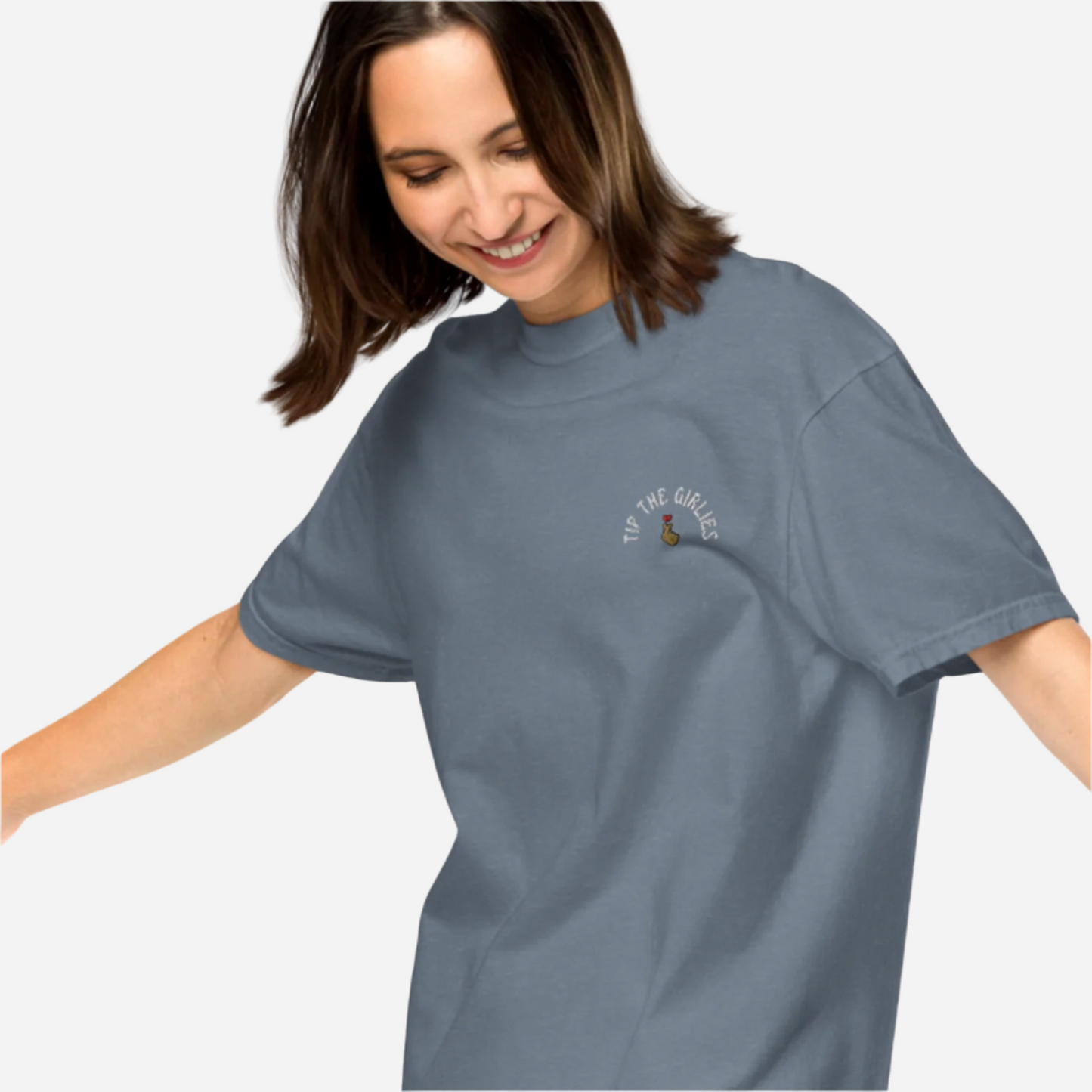 Woman in front of a white background wearing a loose blue tshirt with the words "Tip the Girlies" embroidered on the left chest area.