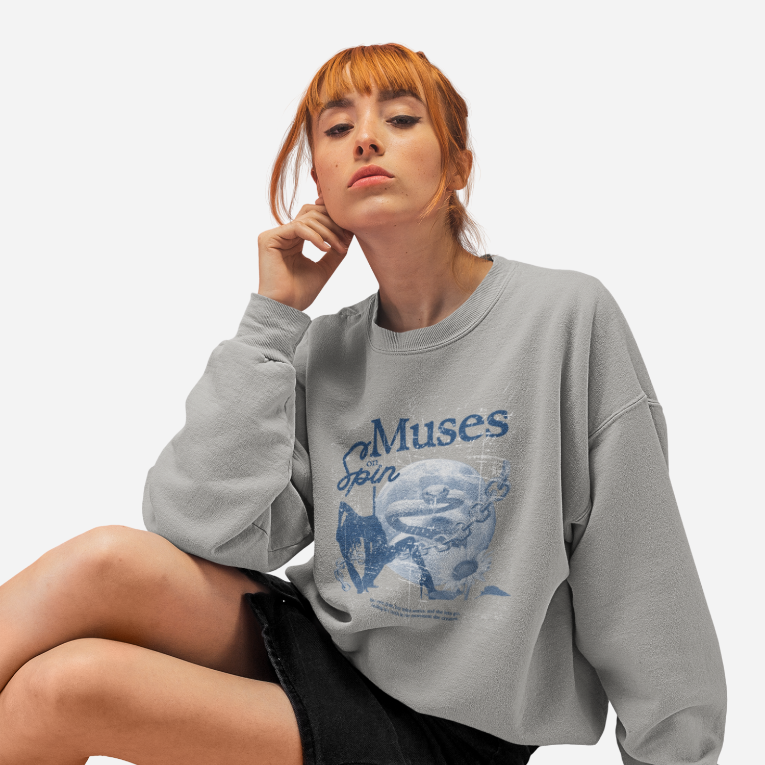 Woman sitting in front of a white background, wearing a grey sweater with a blue graphic that includes a dancer, a moon, a snake, a chain, a flower and the text 