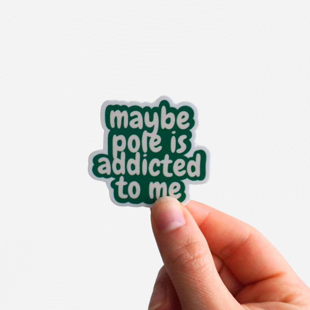 Hand holding a green sticker with text "Maybe Pole is Addicted to Me" on a white background