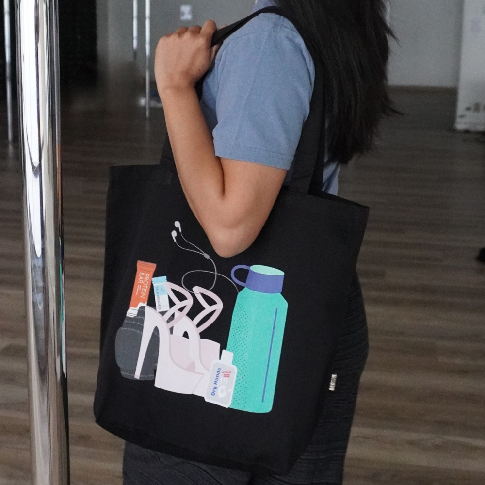 Woman in a pole studio holding a black tote bag with graphic of pole essentials on it - water bottle, heels, knee pads, dry hands and headphones.