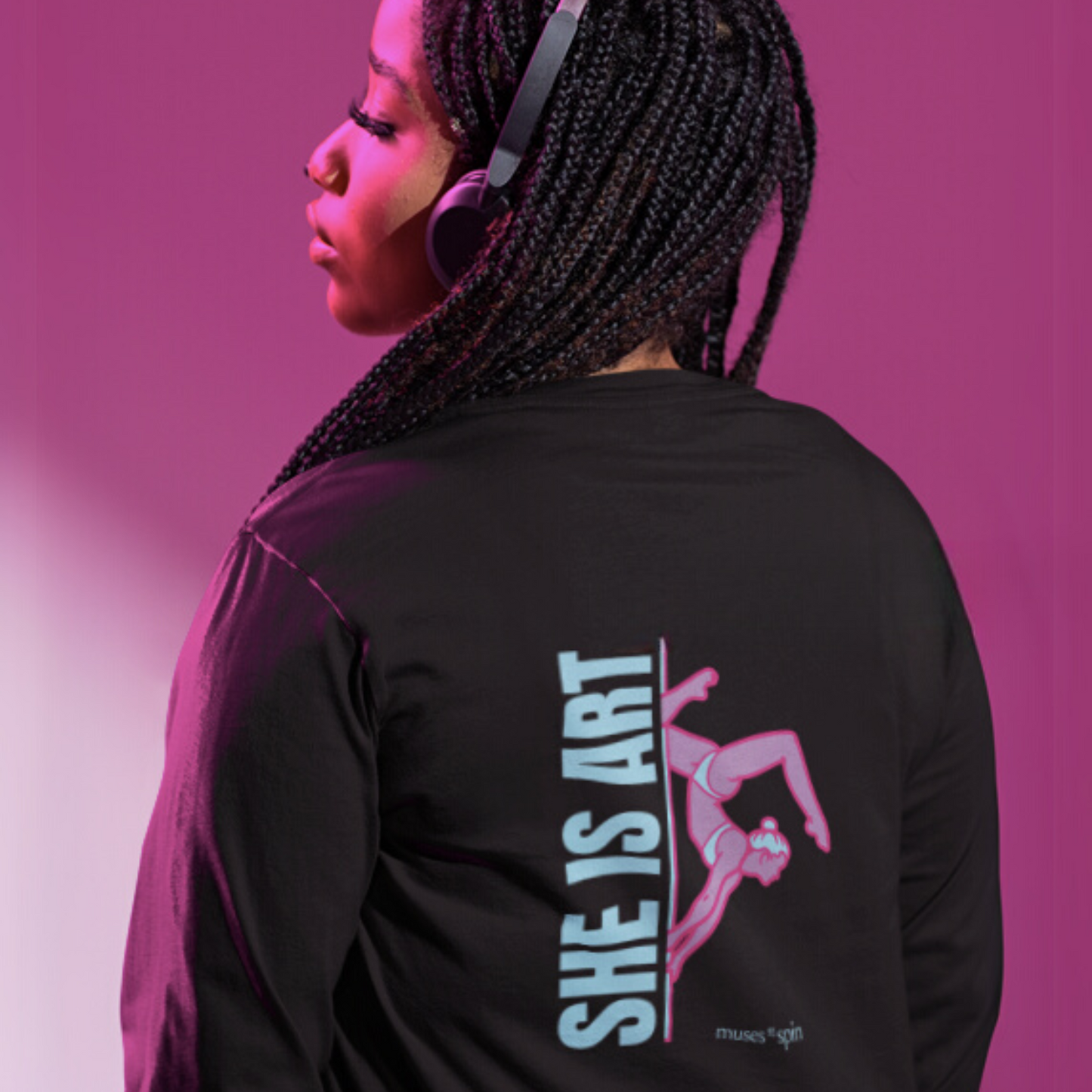 Woman facing a pink background wearing a black long-sleeve shirt with a design saying "She is Art" in light blue and showcasing a pink pole dancer doing an inversion on a pole