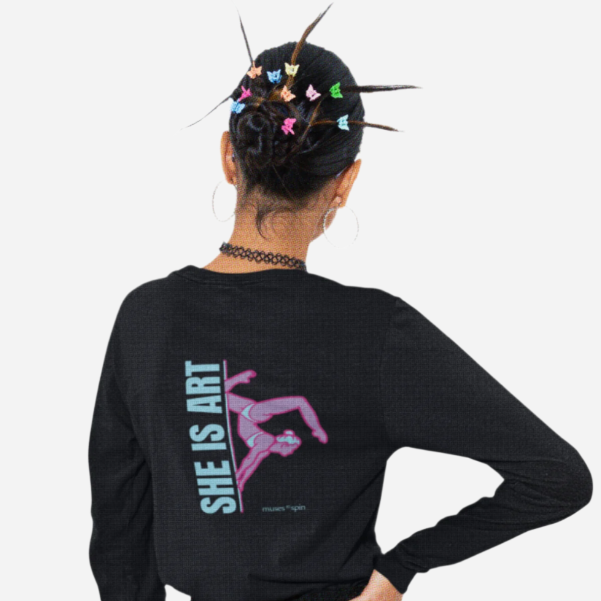 Woman facing a white background wearing a black long-sleeve shirt with a design saying "She is Art" in light blue and showcasing a pink pole dancer doing an inversion on a pole