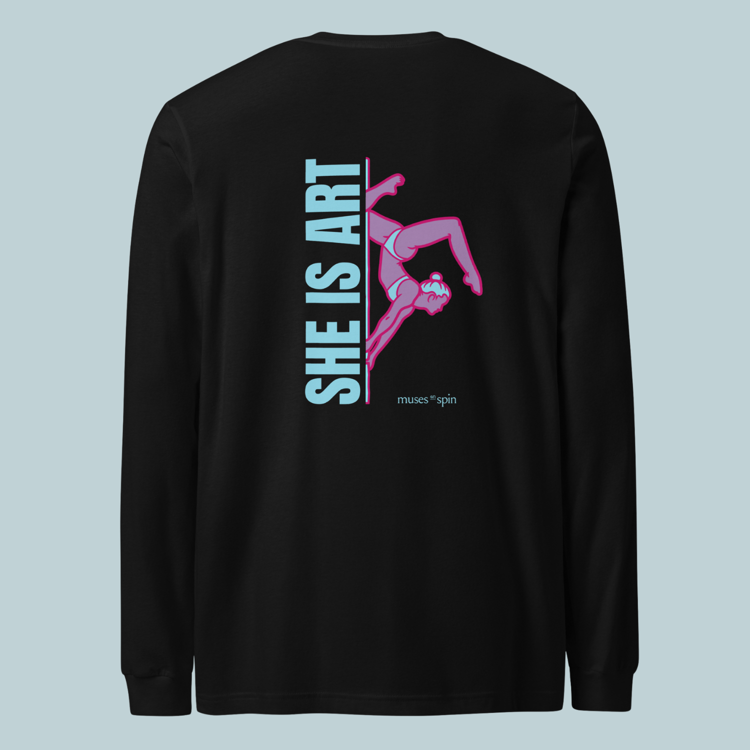 Black long-sleeve shirt with a design saying "She is Art" in light blue and showcasing a pink pole dancer doing an inversion on a pole. The shirt is laid out in front of a light blue background.