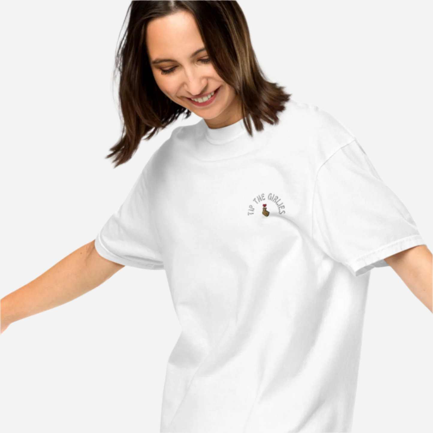 Woman in front of a white background wearing a loose white tshirt with the words "Tip the Girlies" embroidered on the left chest area.