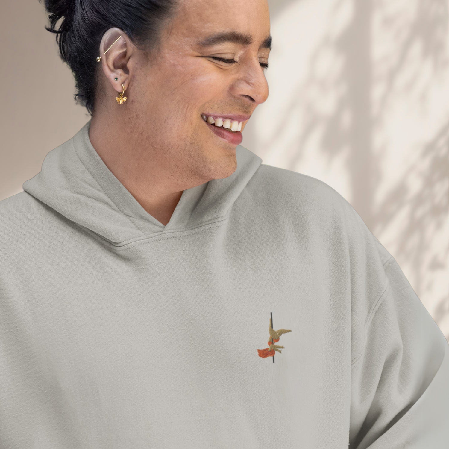Person smiling, wearing a grey hoody with a small pole dancer embroidered on the left chest area. 