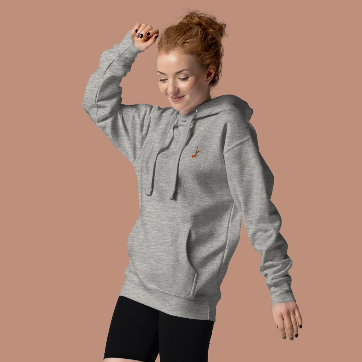 Woman in front of a beige backgroud wearing a grey sweater with a small pole dancer embroidered on the left chest area.