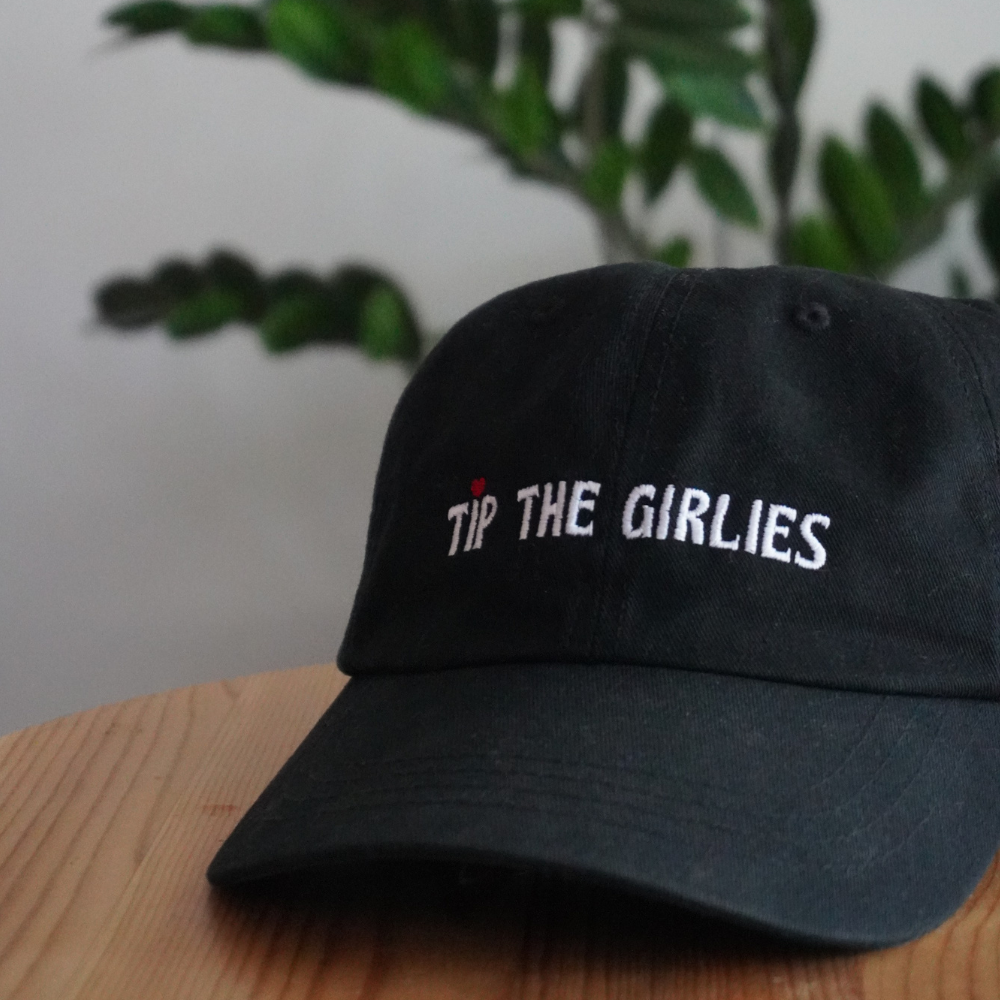 Black cap with 'Tip the Girlies' text on a wooden surface with a plant in the background