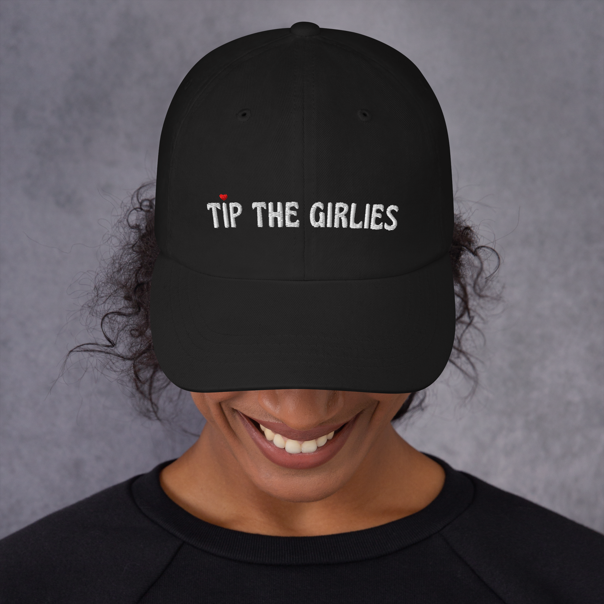 Woman wearing a black cap with the words "Tip the Girlies" embroidered in white letters across the front. The first "I" is topped with a red heart.