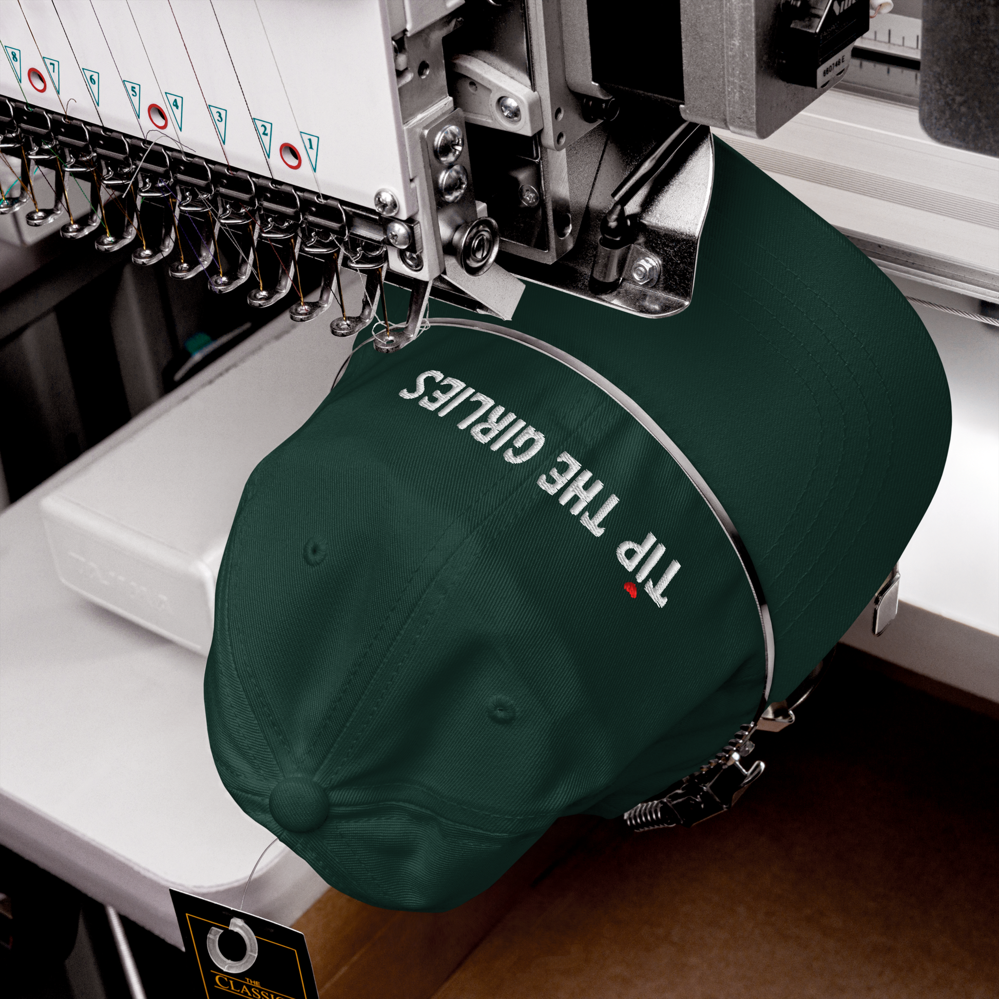 Green cap in the process of being embroidered. The cap has the words "Tip the Girlies" embroidered in white letters across the front. The first "I" is topped with a red heart.