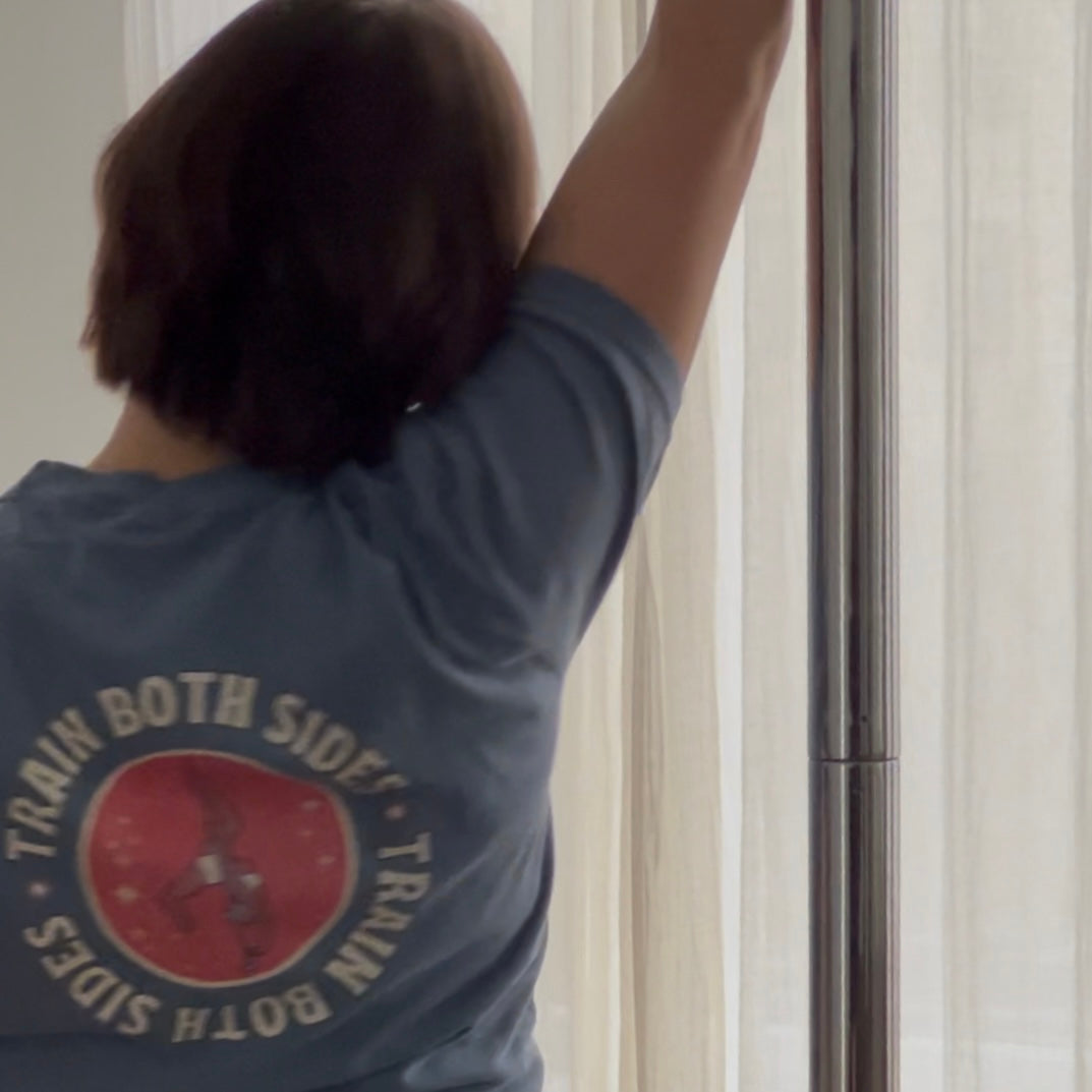 Close-up of a woman pole dancing, wearing a blue shirt with a "Train Both Sides" graphic.