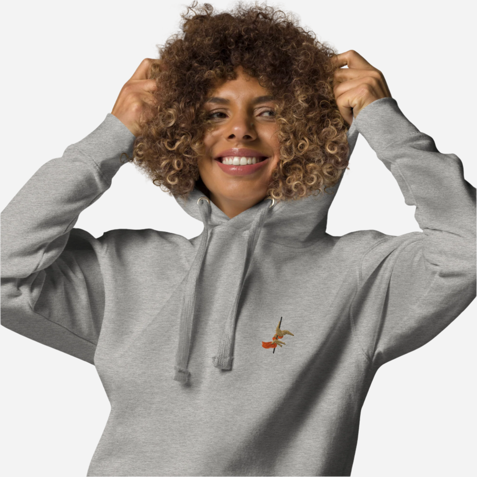 Woman in front of a white background wearing a grey hoodie with a small embroidery of a pole dancer doing an inversion