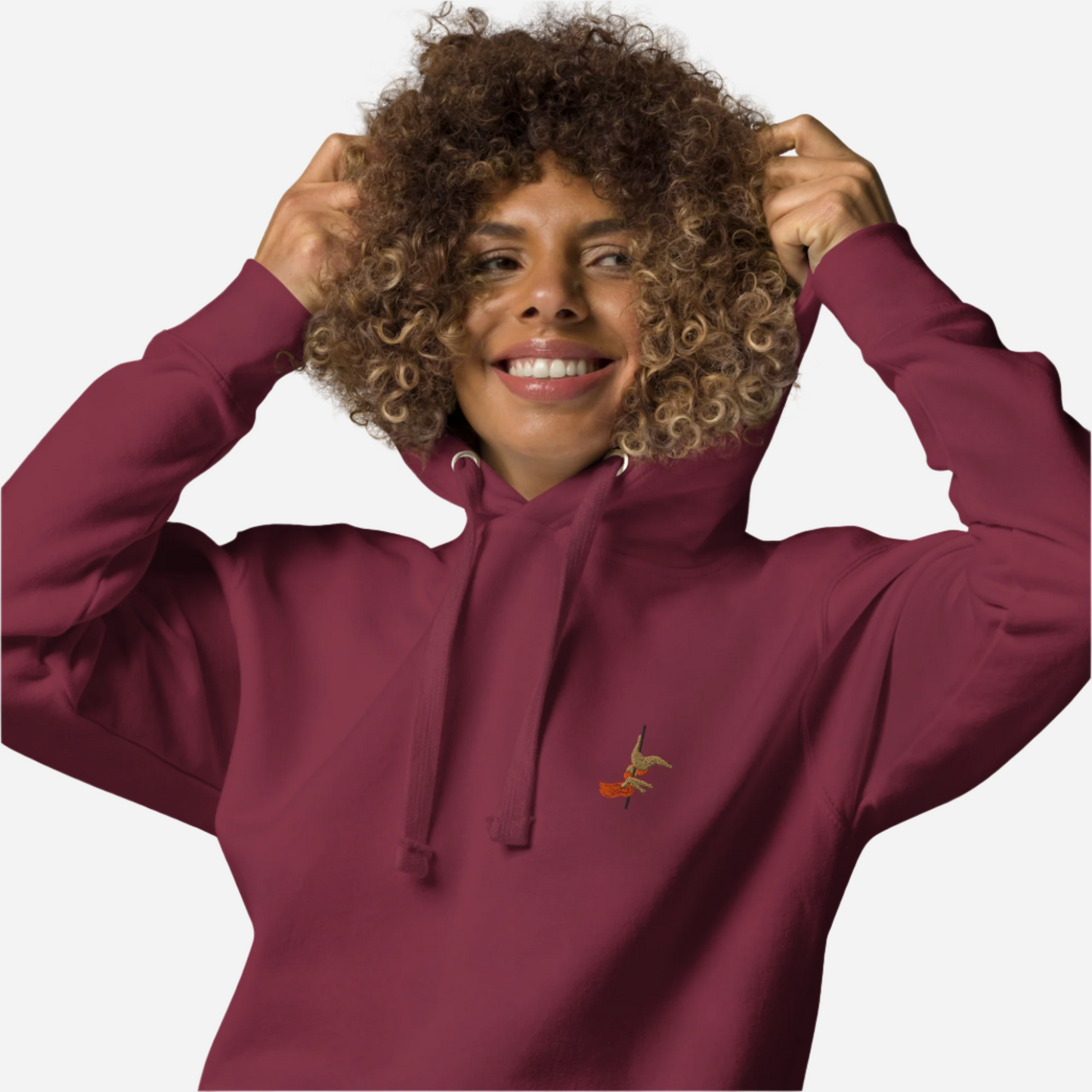 Woman in front of a white background wearing a maroon hoodie with a small embroidery of a pole dancer doing an inversion